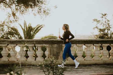A woman jogs on a path in Malta. She wears a black top and blue pants. There are trees and a railing beside her. The sun shines in the morning sky.の写真素材