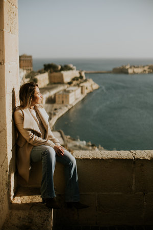 A woman relaxes on a stone ledge, gazing out at the tranquil sea and historic landscape under a clear sky.の写真素材