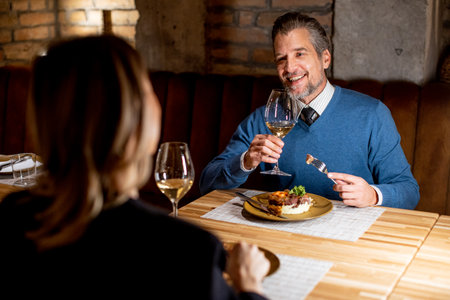A man and a woman sit at a table in a restaurant. They smile and raise their glasses of wine while enjoying dinner. The setting is warm with brick walls and soft lighting.の写真素材