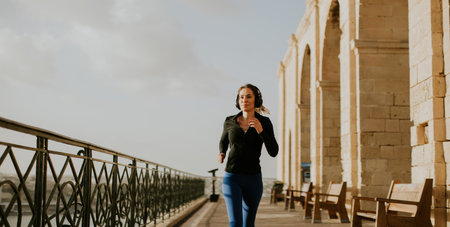 A woman jogs along a path by historic buildings in Malta. She wears headphones and exercises outdoors. The sun shines down as she enjoys her workout near the coastline.の写真素材