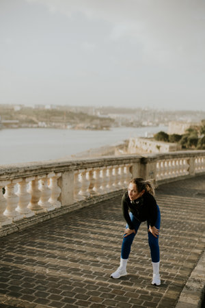 A woman is taking a break from her workout near the coastline in Malta. She is stretching after running on a stone path. The scenery includes water and buildings in the background.の写真素材