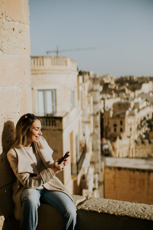 A woman is sitting on a stone wall while looking at her phone. She is in a sunny area of Malta, with buildings and distant views behind her. The scene shows urban life in the city.の写真素材