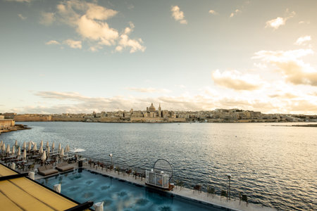 Soft sunlight illuminates the historic Valletta skyline, reflecting on calm waters as visitors enjoy the serene atmosphere along Sliema's coast.の写真素材
