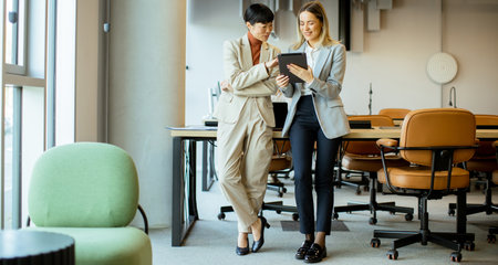 Two women in professional attire engage in a conversation while looking at a tablet. They stand in a bright, contemporary office with stylish furniture and large windows.の写真素材