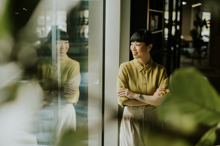 A woman with a short hairstyle stands by a large window in a contemporary indoor setting. She has a...