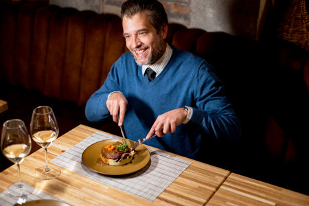A man sits at a wooden table in a restaurant, smiling while cutting his food. He holds a knife and...