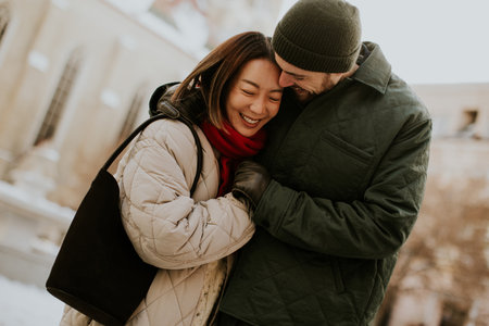 A happy Korean woman and Caucasian man embrace on a snowy day in Europe. The woman is smiling. They are wearing winter clothes, and are outdoors near a building.の写真素材