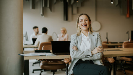 A cheerful businesswoman sits in a stylish office, enjoying a moment of joy while her colleagues...