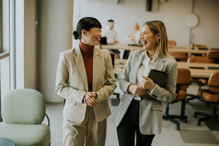 Two women in business attire are smiling and laughing while walking through a modern office space. The atmosphere is relaxed, with colleagues in the background engaged in work.の写真素材