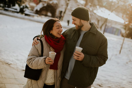 A smiling Korean woman and Caucasian man are walking together outside in the winter. They are both...