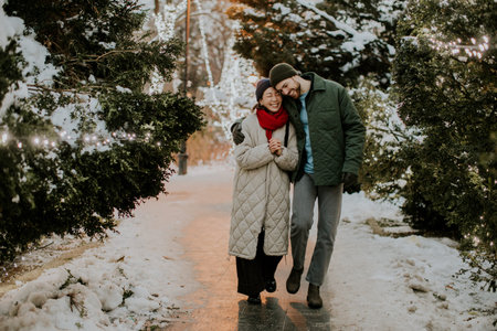 A Korean woman and a Caucasian man walk hand in hand along a snowy path in a park. They smile and share a moment together, surrounded by trees decorated with lights on a winter evening.の写真素材