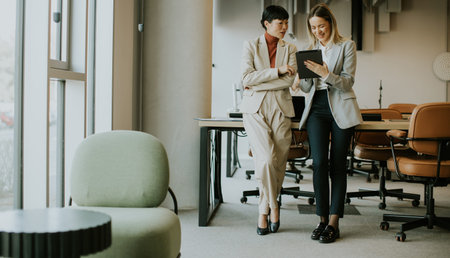 Two professional women stand in a contemporary office, looking at a tablet. They share insights and ideas in a collaborative environment filled with stylish furniture and natural light.の写真素材
