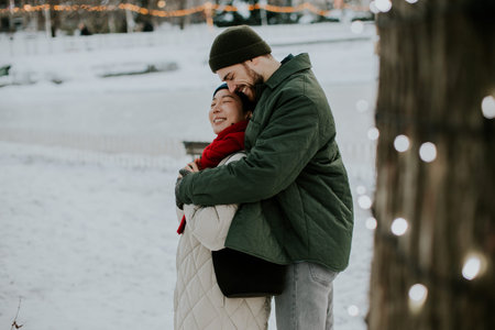 A Korean woman and a Caucasian man smile warmly at each other in a snowy park. They stand close...