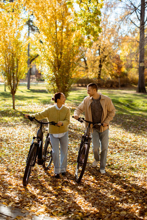 In a vibrant park filled with autumn leaves, a Korean woman and a Caucasian man enjoy a cheerful bike stroll, exchanging smiles and laughter.の写真素材
