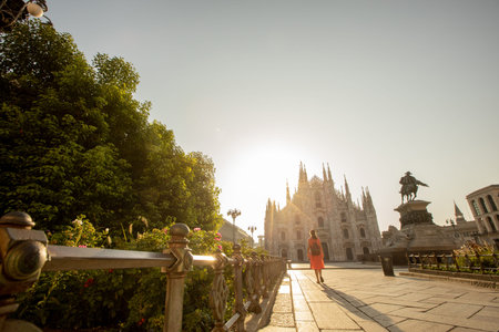 A young woman walks gracefully near the iconic Duomo cathedral in Milan, illuminated by the soft light of sunset, surrounded by vibrant greenery.の写真素材