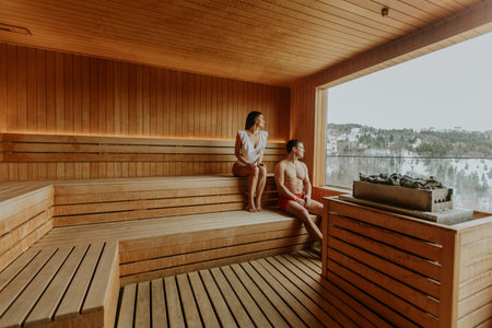 Two individuals sit comfortably in a wooden sauna, soaking in warmth while gazing at the frosty scenery outside.の写真素材