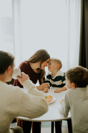 A family enjoys breakfast at a round table. Two children share smiles with their mother. One adult drinks coffee while another child watches. Sunlight comes through the window.の写真素材