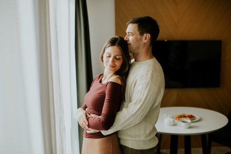 A man and woman share a close moment in an indoor space. They are standing by a window in the...