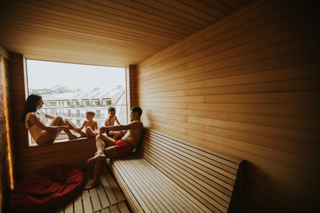 A family sits in a wooden sauna. Two adults and two children relax inside the warm space. They talk...