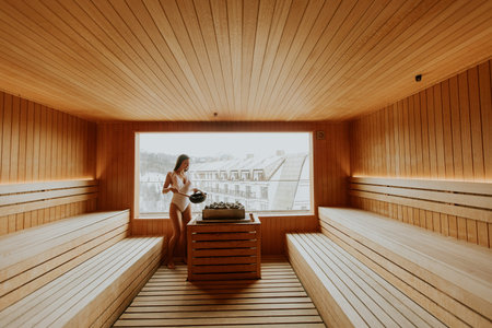 A woman stands in a sauna holding a bowl. She looks out through a large window at a winter landscape. The wooden interior creates a warm atmosphere for relaxation.の写真素材