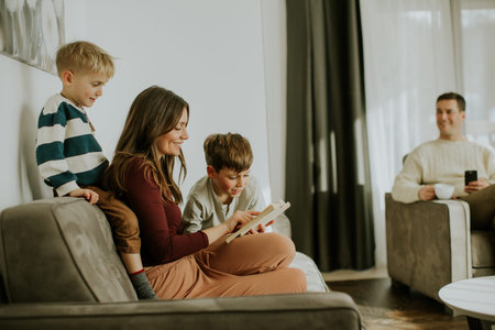 A family spends time in their living room. A mother reads a book to her two young boys. One boy sits on her lap, while the father sits nearby holding a cup.の写真素材