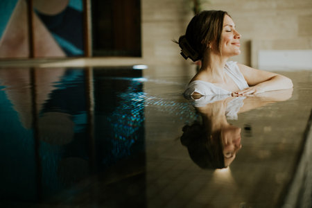 A woman relaxes in a pool at an indoor spa. She sits with her eyes closed and smiles gently. The...