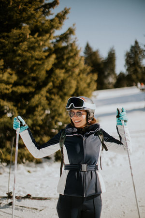 A woman stands on a snowy slope holding ski poles. She wears ski gear, a helmet, and goggles. Trees are in the background under a clear sky. It is daytime and she looks happy.の写真素材