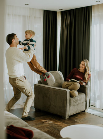 A man lifts a young boy in a living room. The woman sits on a couch nearby. The room has large windows with curtains. The family enjoys time together in the cozy space.の写真素材