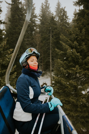A woman sits on a ski lift wearing a ski suit and goggles. She is surrounded by tall trees covered in snow. The weather looks cloudy and cool as she enjoys the ride up the mountain.の写真素材