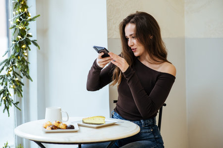 A woman sits at a table in a cafe, using her smartphone. She has a dessert on the table and a cup...