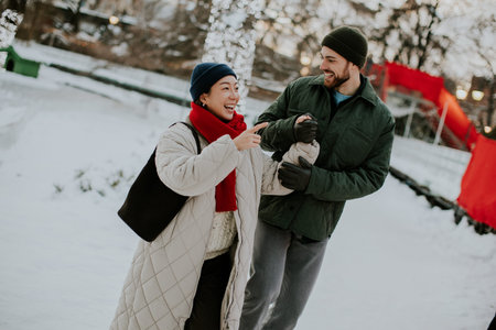 A couple walks hand in hand through a snowy park filled with trees and holiday lights. They are...