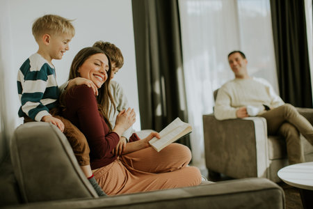 A woman sits on a couch reading a book while two children smile and enjoy the moment. A man sits...