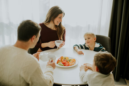 A family sits around a table in their home enjoying breakfast. The parents drink coffee while the...
