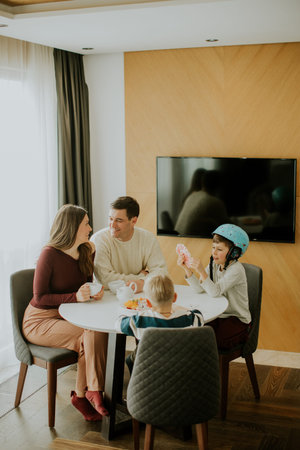 Four family members enjoy snacks and drinks around a round table in their living room. They talk and...