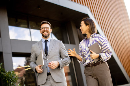 A Korean woman and a Caucasian man walk outside a modern building. They talk and smile while holding folders. The sun shines on them, creating a bright atmosphere.の写真素材