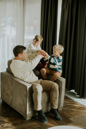 A man sits on a chair with two boys around him. They appear to be playing a hand game. The setting is a warm living room with natural light coming from the windows.の写真素材