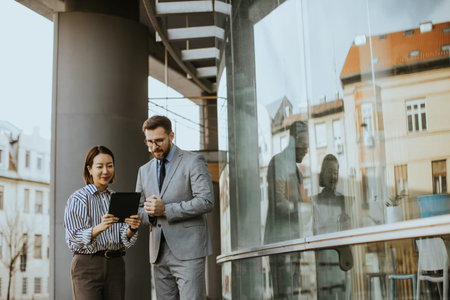 A Korean woman shows a tablet to a Caucasian man on a busy city street. They stand near a glass...