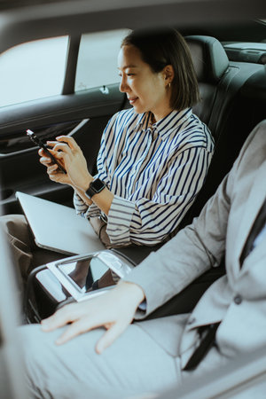 A woman sits in a car focused on her phone while a man sits beside her. They are both dressed in...