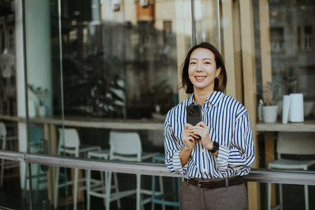 A Korean woman stands outside a cafe holding a smartphone. She smiles at the camera while leaning against the glass wall. The setting shows urban life during daytime.の写真素材