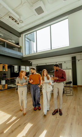 Four people walk together in a modern office space. They are smiling and talking with each other. Bright sunlight comes through the large windows. The mood is friendly and relaxed.