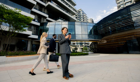 Two individuals walk past a person standing in a business suit using a smartphone. This scene takes place near modern office buildings and trees in the background.