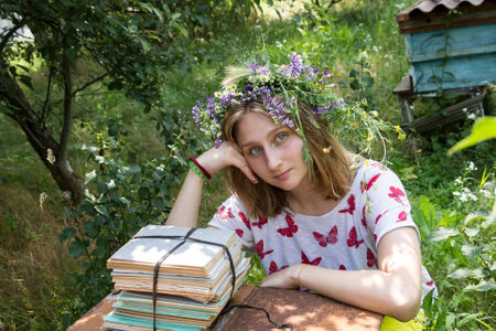 young girl student in a wreath of field herbs next to a stack of notebooks on vacation in natureの写真素材