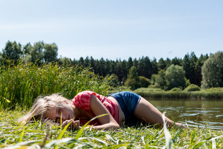 a young girl in blue shorts and a pink t-shirt lies in the grass on the beach near a forest lakeの写真素材