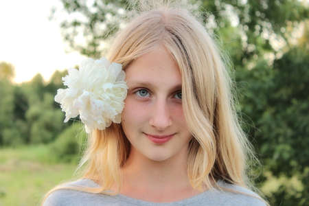 young student girl in a T-shirt with a white peony flower in her hair stands in natureの写真素材