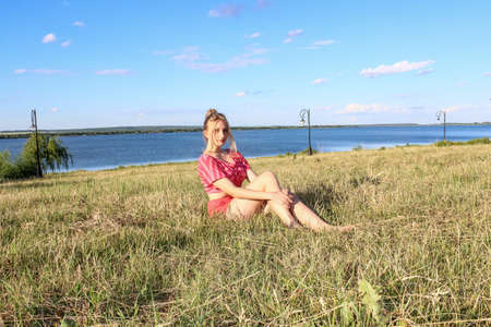 a young girl student in shorts and a T-shirt sits in the grass on the embankment by the riverの写真素材