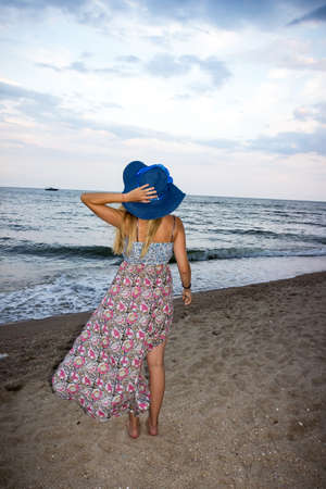 a young girl in a blue hat and a sundress stands in the evening on a sandy beach and looks at the seaの写真素材