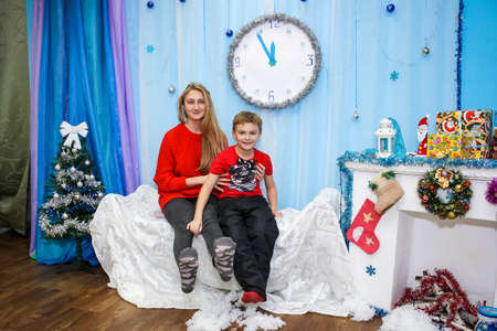 a young girl and a boy are sitting on an ottoman by the Christmas tree with Christmas gifts and artificial snowの写真素材