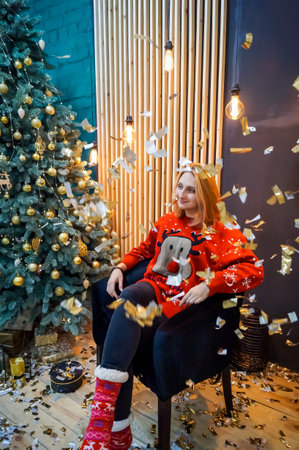 young blonde girl sitting in a chair in a red wreath and in a red New Year's sweater under flying sparklesの写真素材