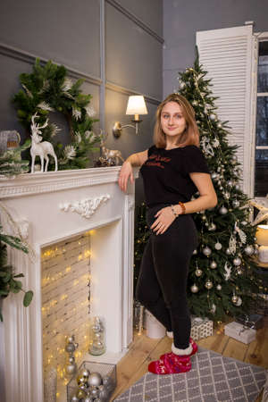 a young blonde girl in a New Year's t-shirt stands by the tree near the fireplace in New Year's decorationsの写真素材