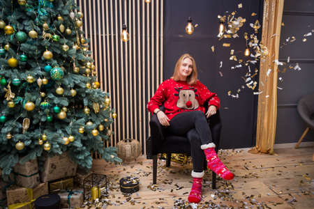 young blonde girl sitting in a chair in a red New Year's sweater under flying sparklesの写真素材